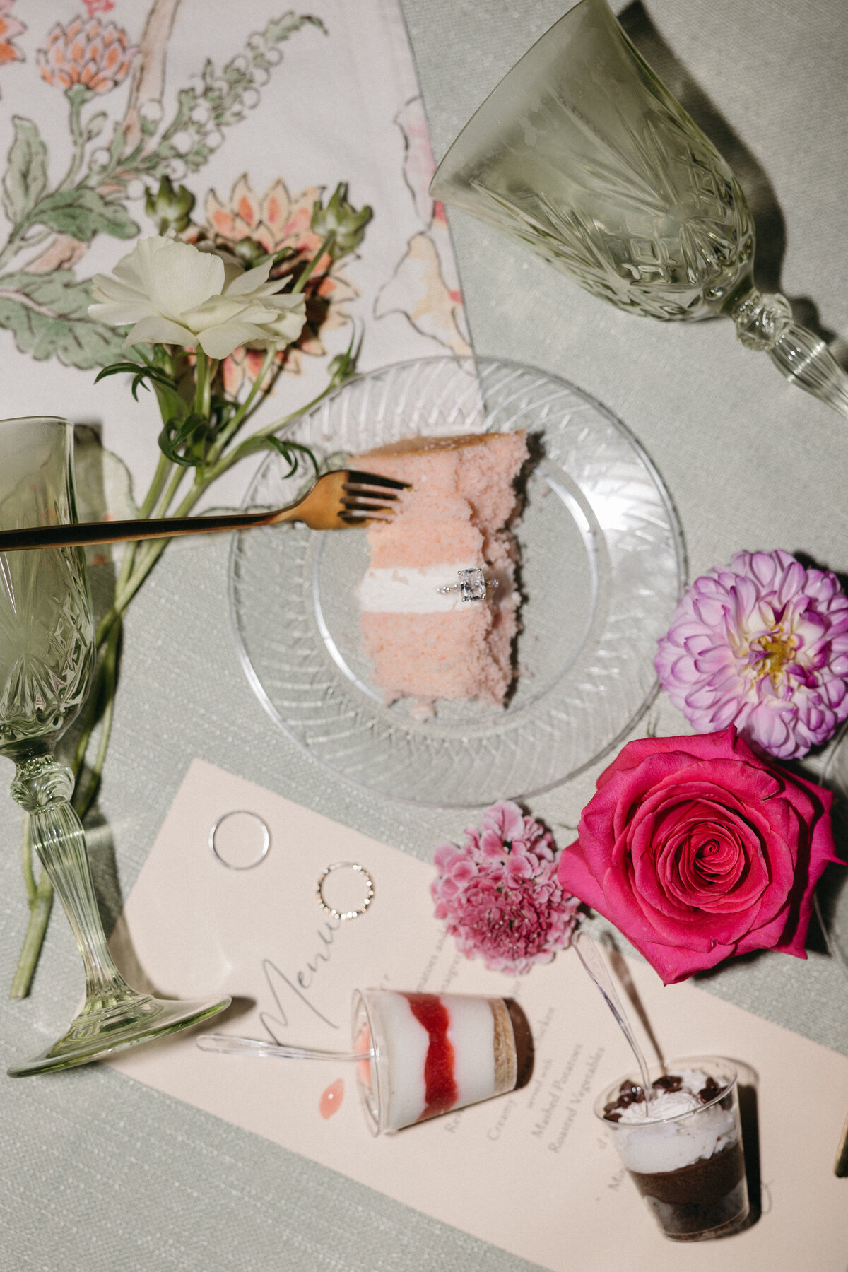 Romantic wedding detail flatlay with pastel flowers, textured linens, vintage glassware, dessert cups, and a slice of pink cake showcasing elegant wedding styling and floral artistry.
