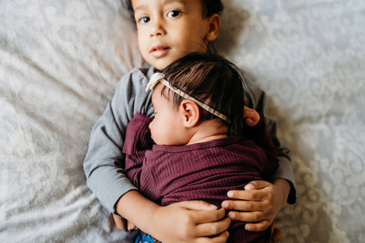 Newborn Photographer, a little baby sleeps on top of her older brother, his arms wrapped around her