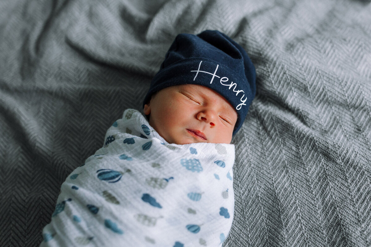 swaddled newborn with hat that says Henry sleeping on hospital bed