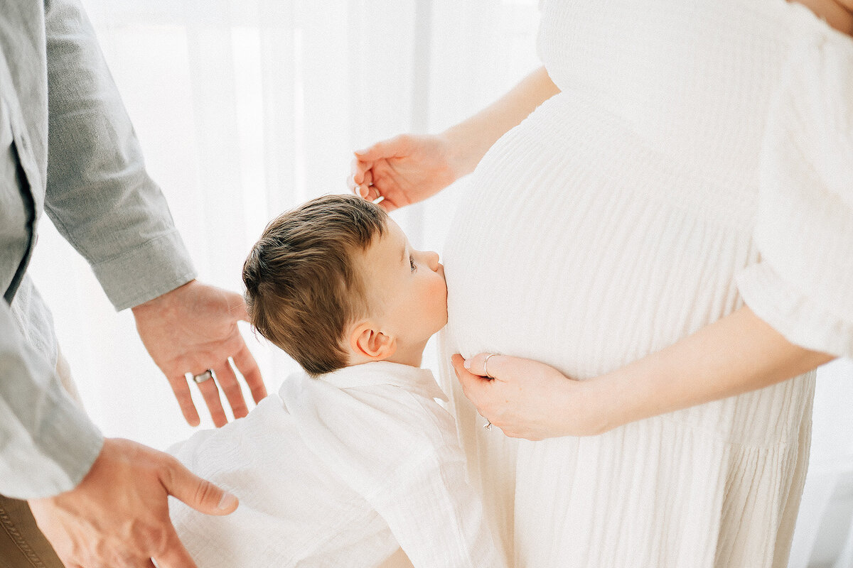 pregnant mother with child leaning in kissing her belly with dad's hands keeping him safe