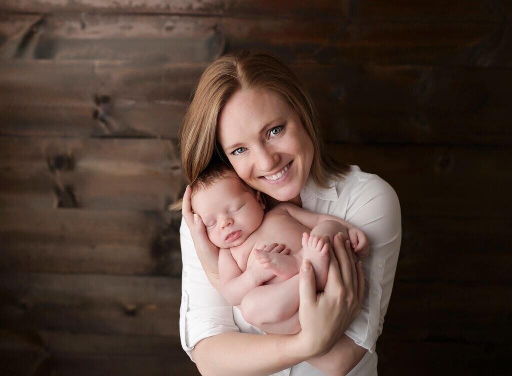 Emily, the photographer holding her newborn son.  She is wearing a white collared shirt in front of a dark wood backdrop.