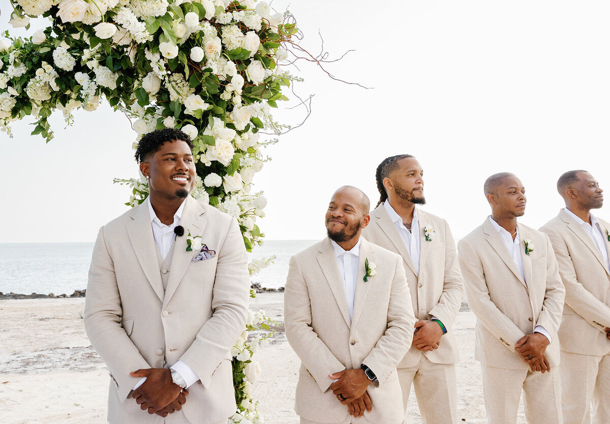 Jonathan’s groomsmen stand in beige suits, smiling as they wait for the bride’s arrival at their stunning beachfront wedding in Table Key, Islamorada. Captured by Claudia Amalia Wedding Photography.