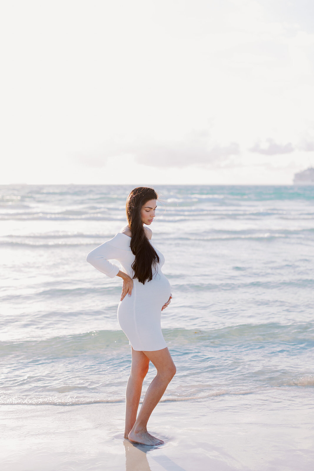 A glowing expectant mother gazes at the horizon, waves gently lapping at her feet.