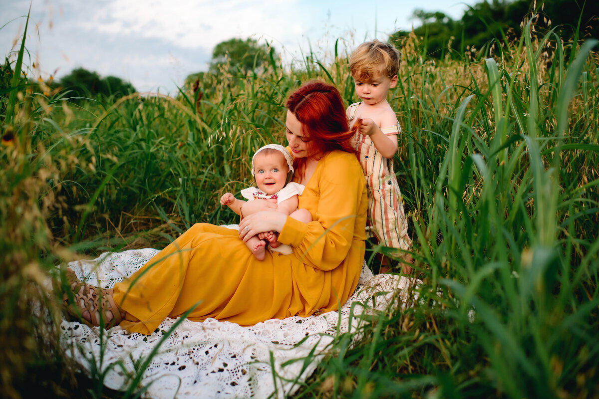 The essence of family photography with this image of an Albuquerque family photographer sitting on a white blanket with her beautiful babies. Dressed in a yellow long dress, she is surrounded by lush green grass, highlighting the serene and joyful moments of family life in Albuquerque.