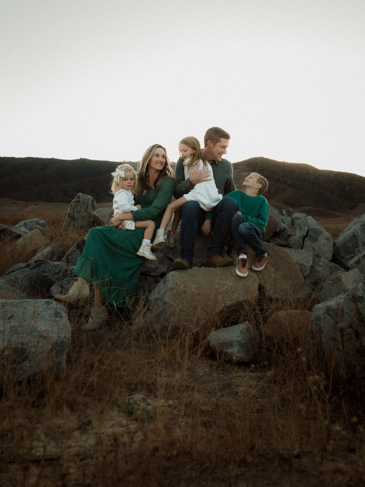 Family of five sitting together on boulders outdoors, laughing and cuddling in the evening light.
