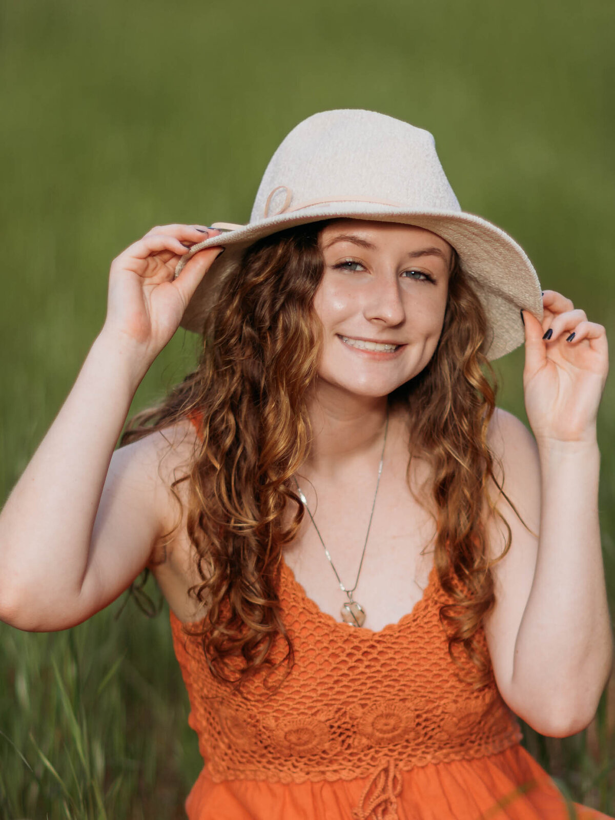 A smiling senior portrait of a girl in her orange dress and wearing a white hat.