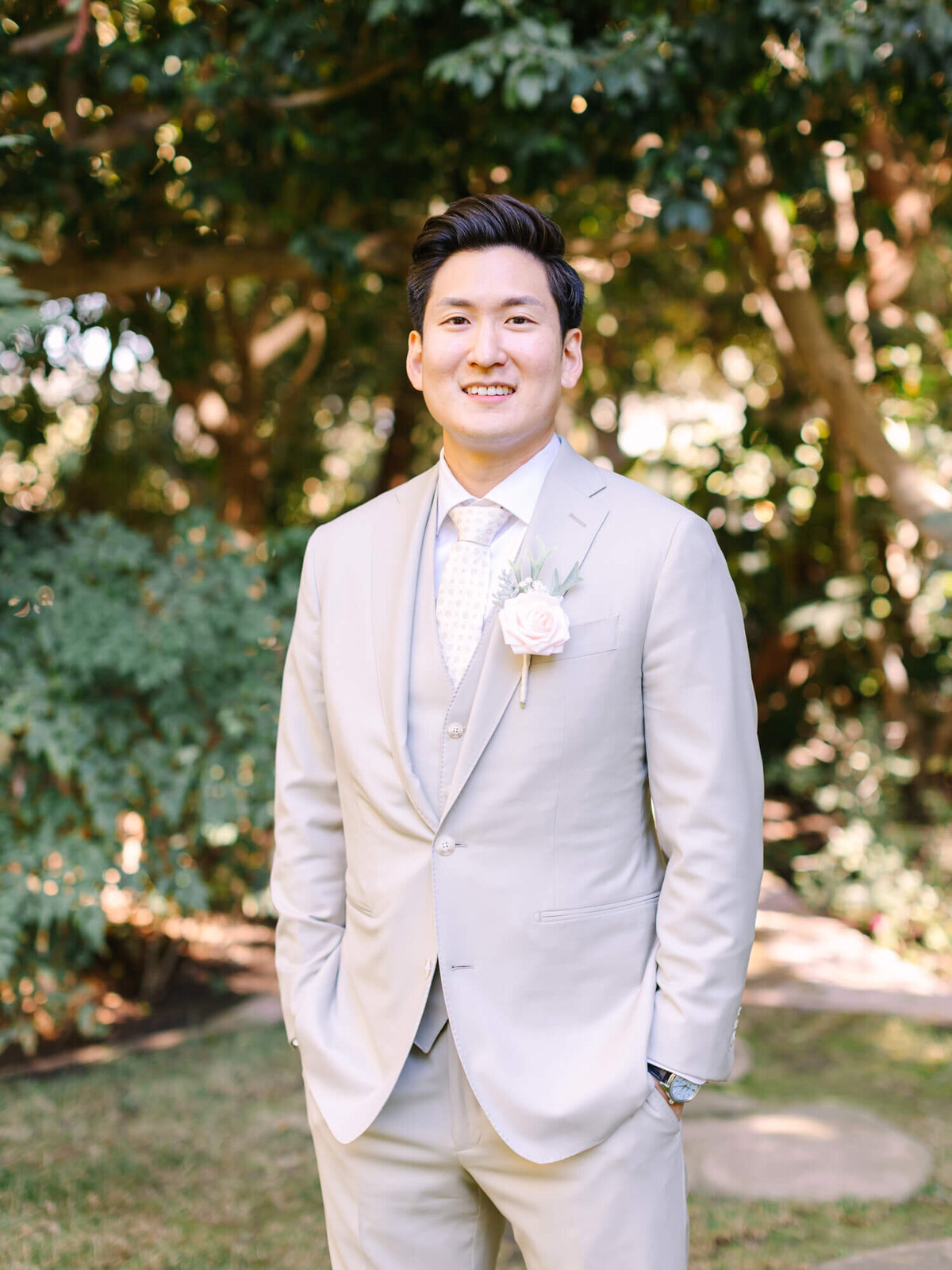 A groom wearing a light beige suit and tie with a white rose boutonniere smiles warmly while standing in a lush garden.