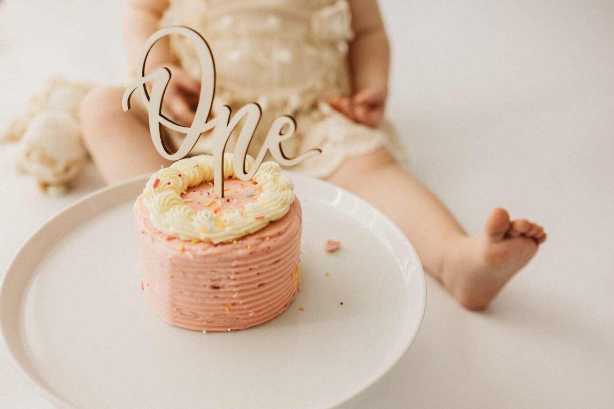 Ruua Hamdan Photography (UK): A small pink and white cake with a "One" topper sits on a plate, with a baby's legs softly blurred in the background.