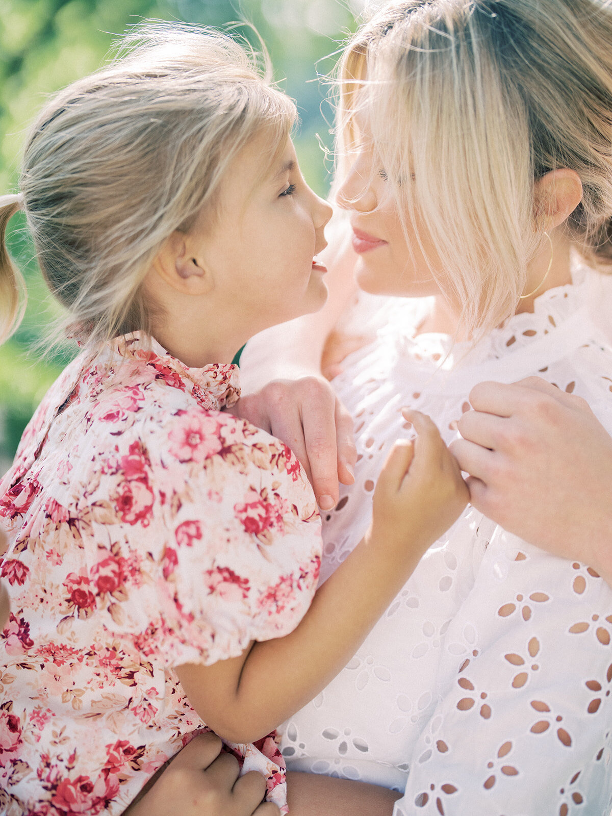 Blonde mother leans into her toddler daughter in pink dress while holding her.