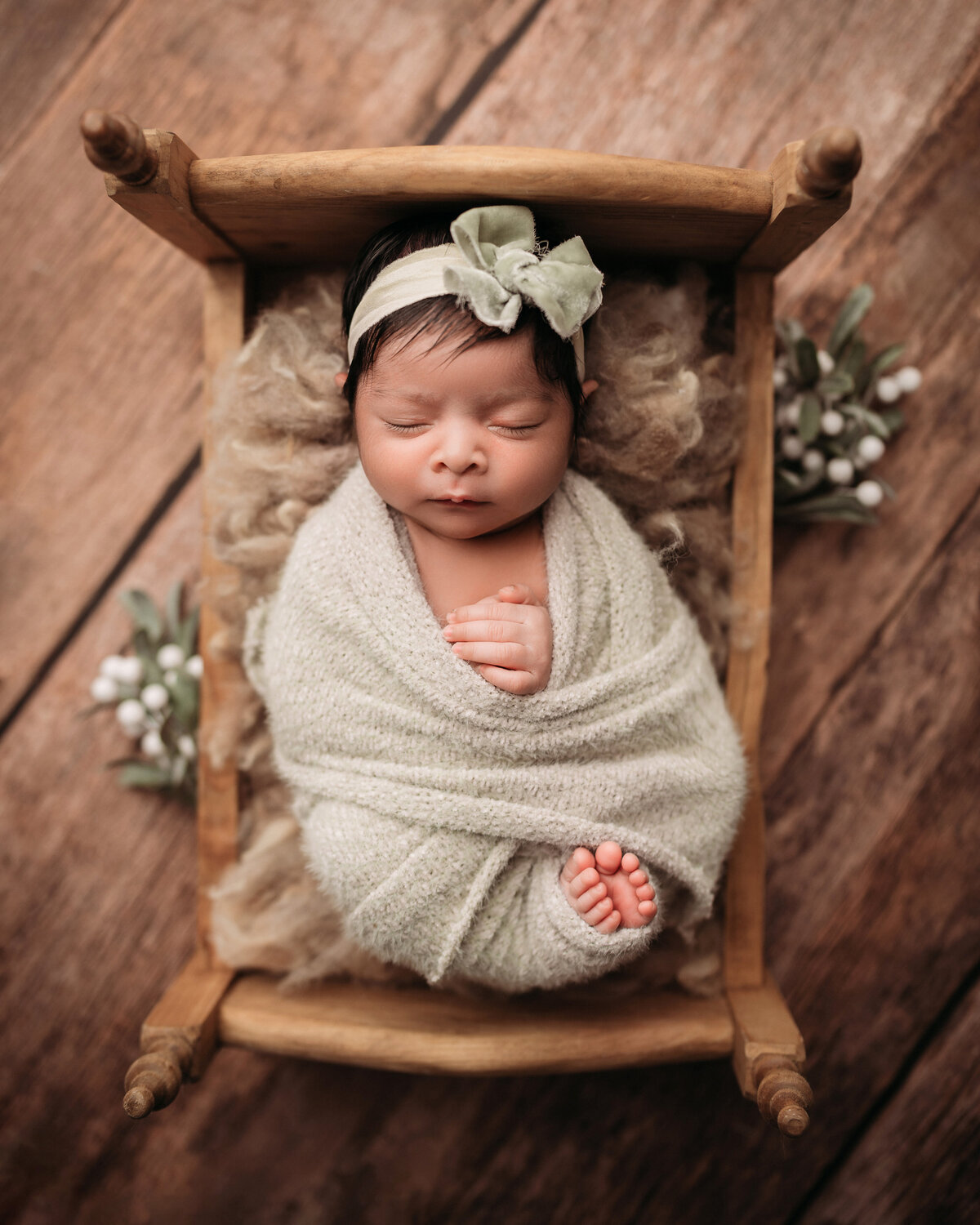 sleeping newborn baby girl with dark hair wearing matching sage green bow and swaddle  and her toes are peeking out while she is posed inside of tiny wooden bed with a wooden backdrop