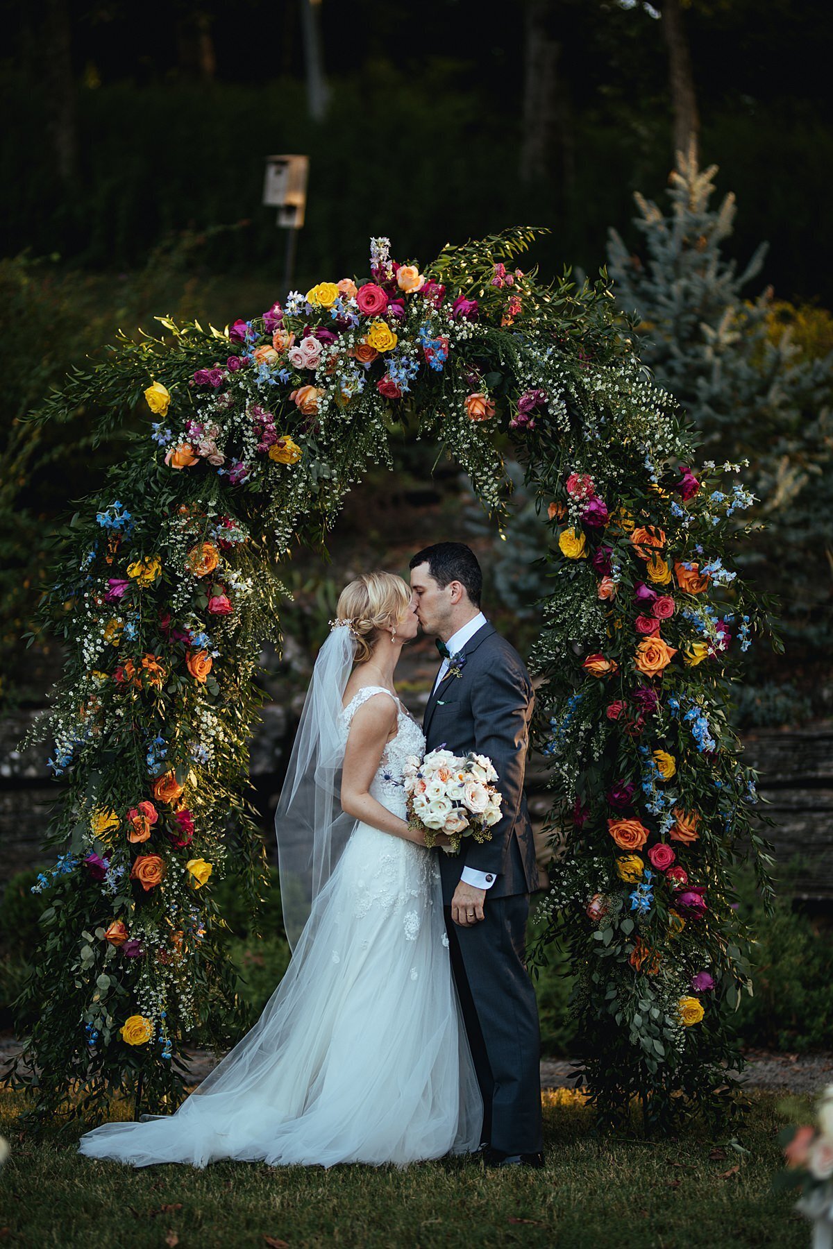 The bride, wearing a long veil and long lace wedding gown kisses the groom in a charcoal gray suit while holding a large white and blush bouquet in front of a brightly colored floral arbor with yellow, hot pink, blue, purple, white, ivory, red and orange flowers at Cheekwood Botanical Garden