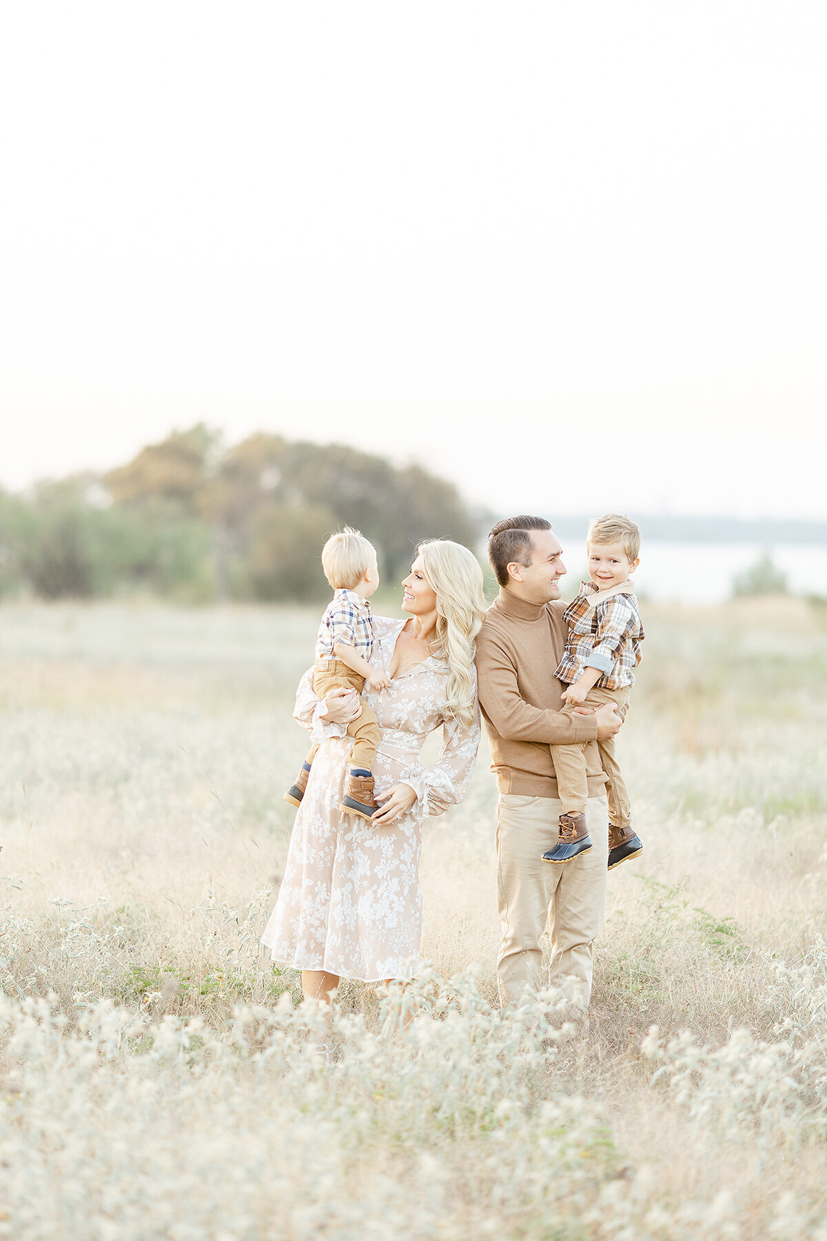Family photo of a mother and father holding their two young boys as they pose for family photos in the middle of an open field at a Dallas park.