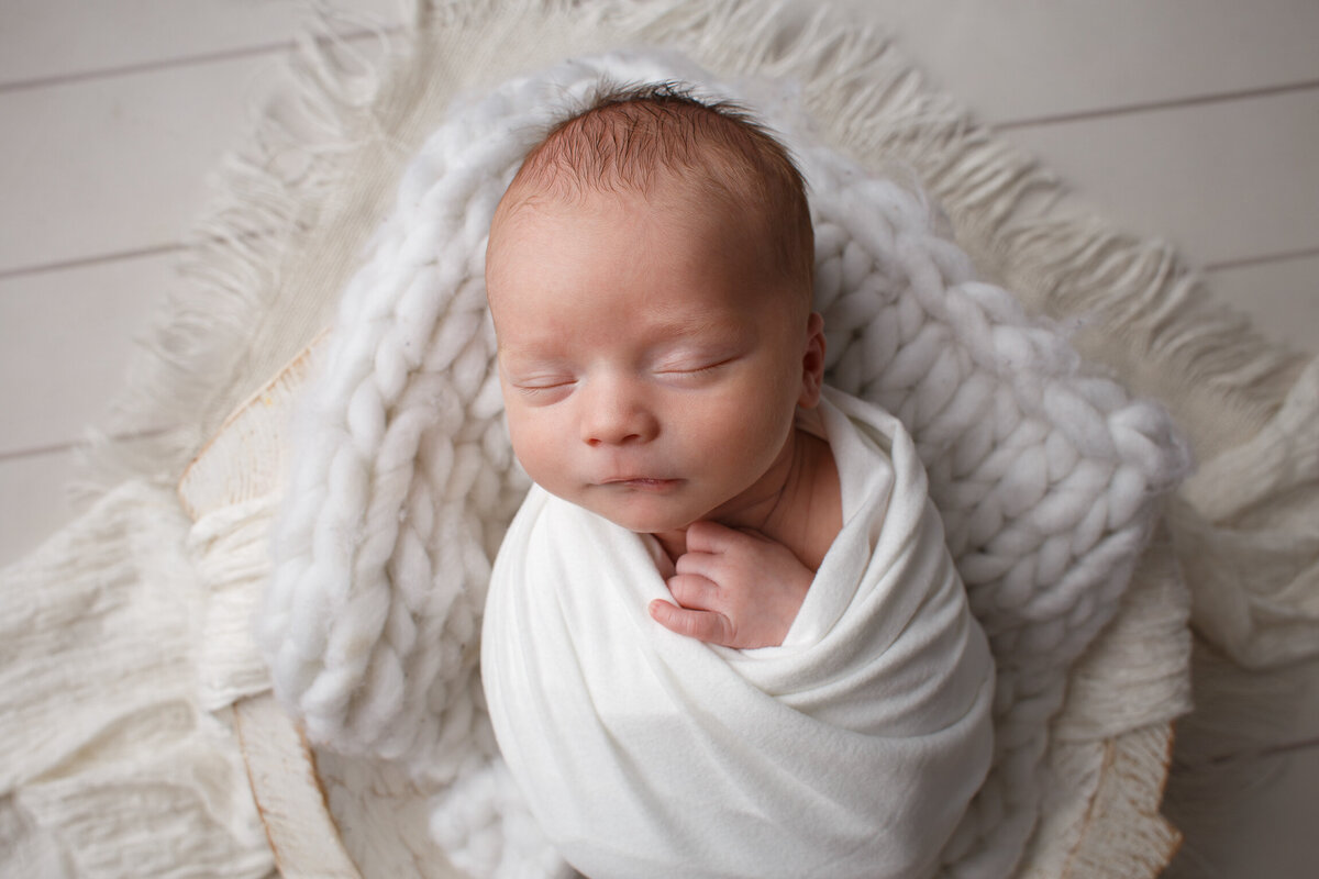 Image of a newborn in a wite wrap laying on a white blanket in a basket