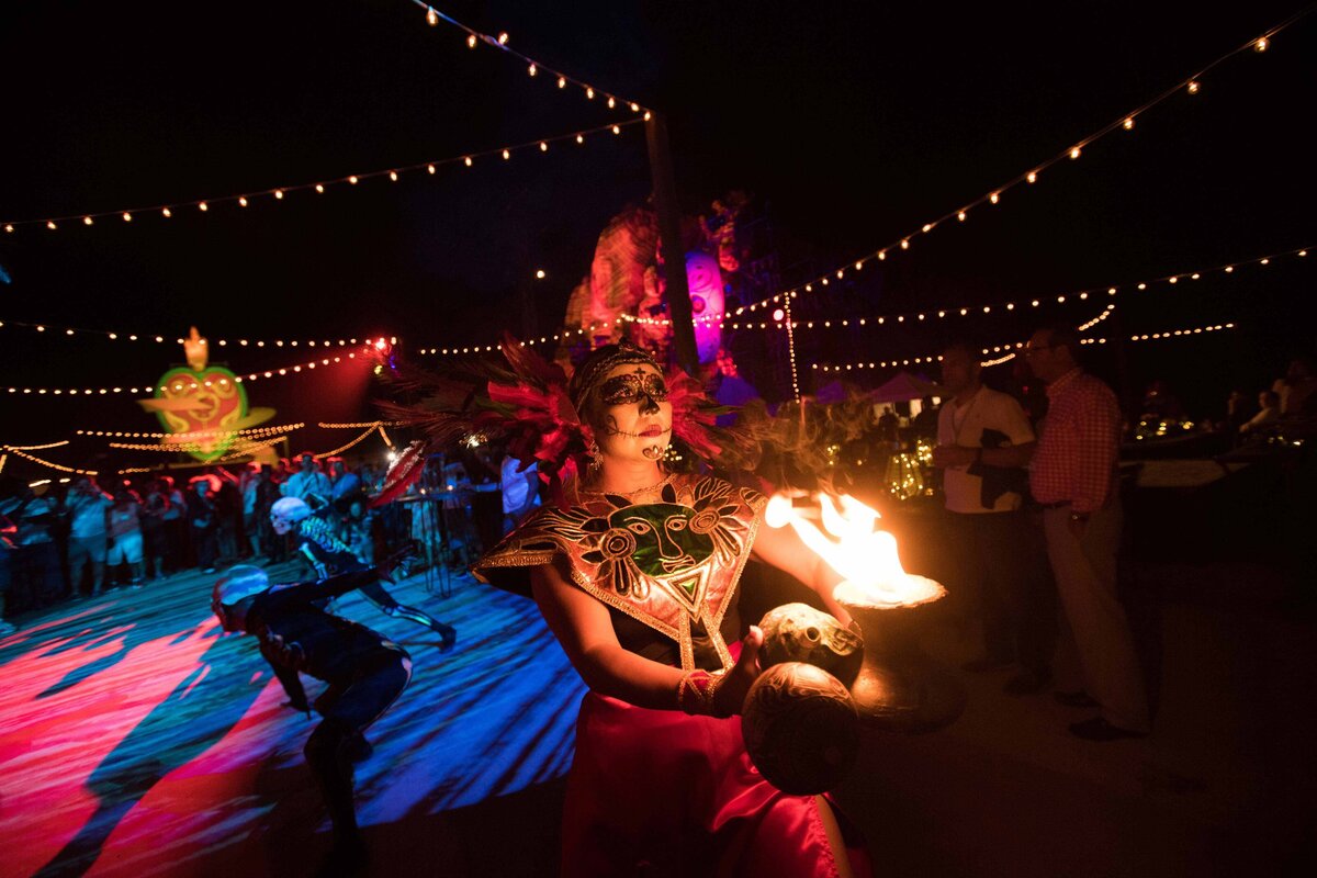 An actor in Cabo with day of the dead face paint and costume walks holding a pot of fire while surrounded by dances and guests.
