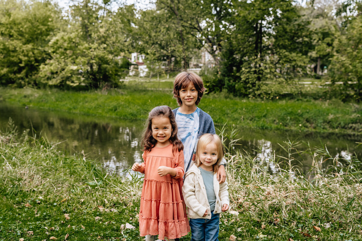 Three kids stand in front of a pond outdoors.