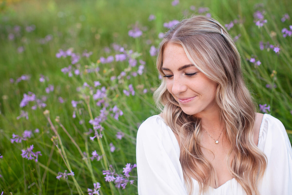 A woman with long, wavy blonde hair and a white dress smiles gently while sitting in a field of purple flowers. Her eyes are closed, and the green grass creates a serene backdrop.