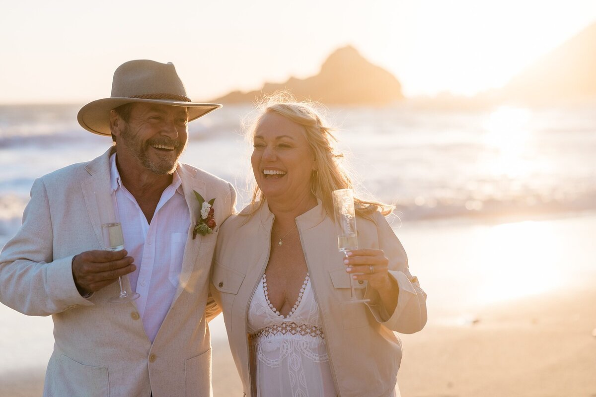 The bride and groom walk along the beach and laugh during sunset. 