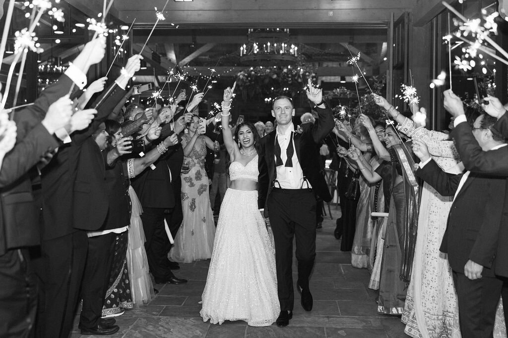 Black and white sparkler exit photo of the couple leaving their reception surrounded by cheering guests.