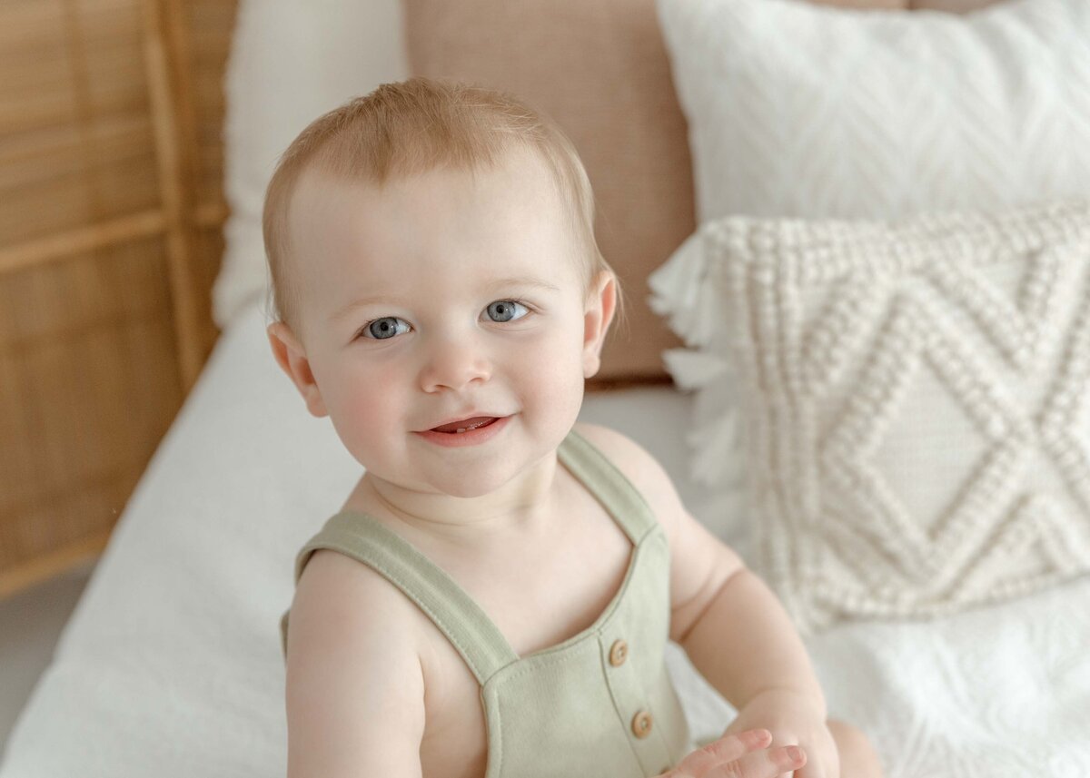 One year old boy sitting on bed smiling dressed in sage overalls