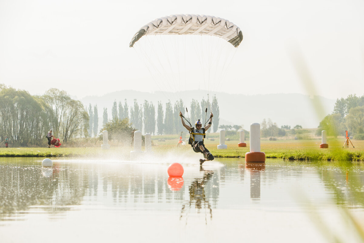 Ein Fallschirmspringer fliegt über das Wasser. Das Bild zeigt einen Fallschirmspringer, der mit einem Fuß im  Wasser streift. Zu sehen ist seine Reflektion und dieUmgebung