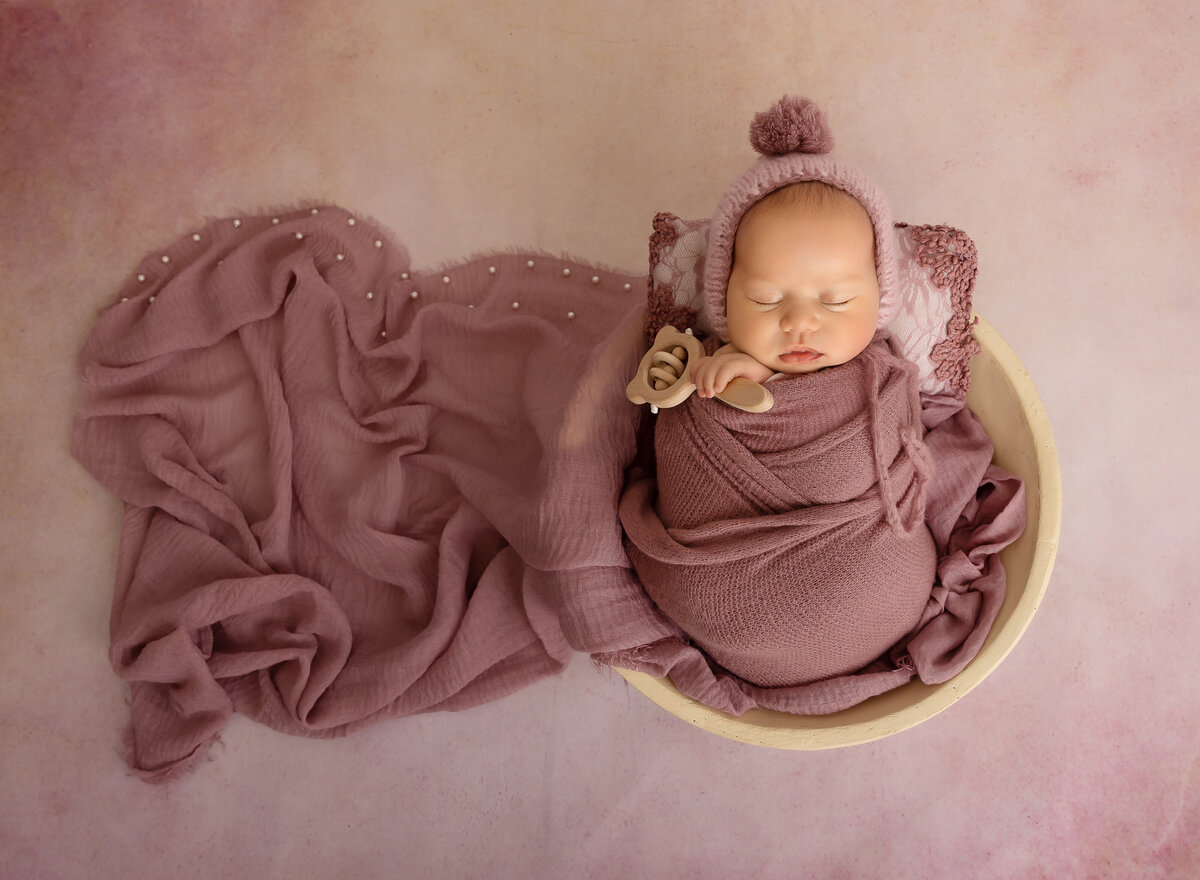 Brooklyn newborn photoshoot. Baby girl is placed in a bowl and wrapped in a mauve swaddle that is ruched over the side of teh bowl. She is sleeping and holding a tiny wooden rattle in one hand. Captured by top Brooklyn newborn photographer Chaya Bornstein