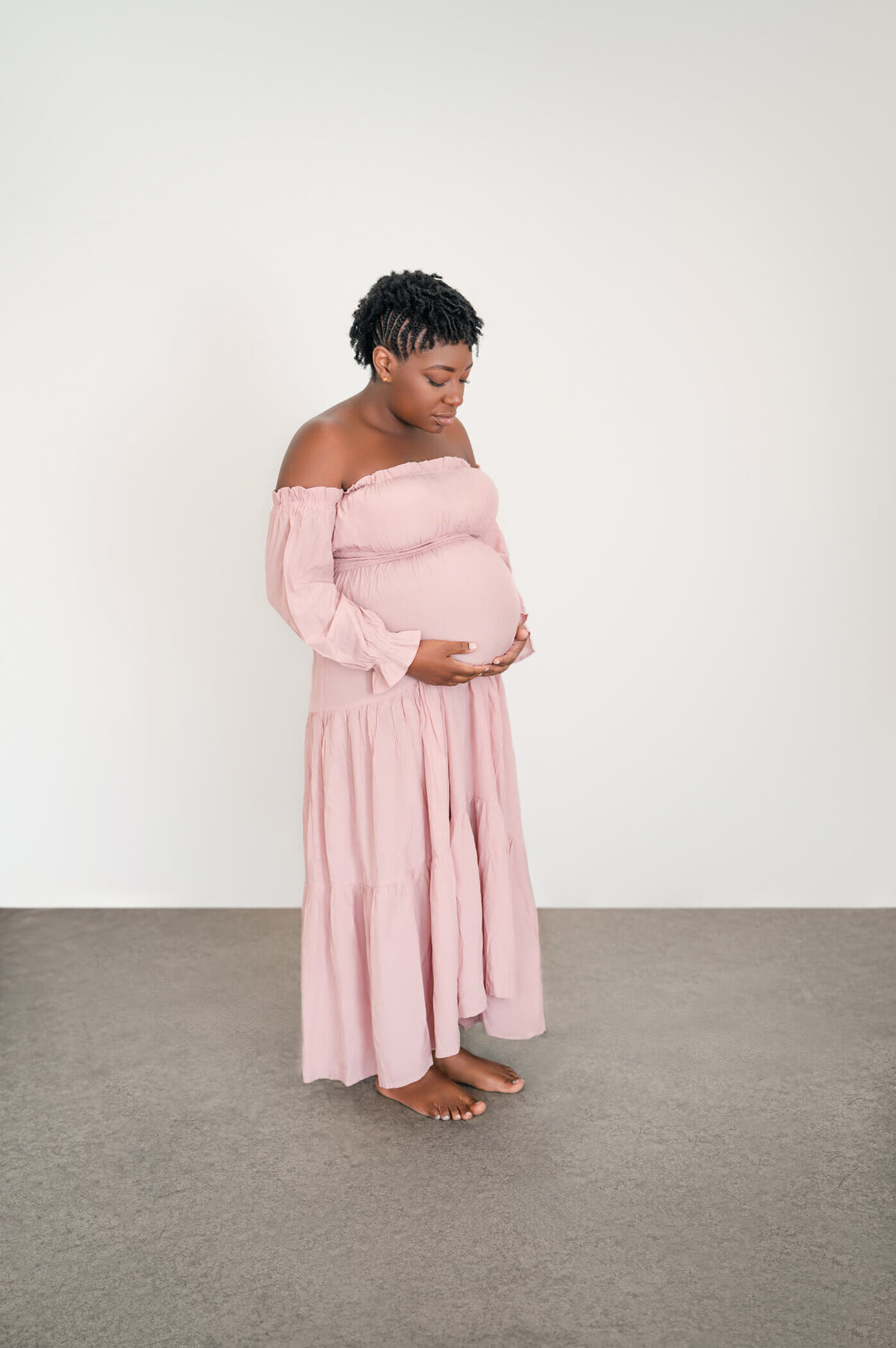 beautiful mama holding her pregnant baby bump in an off shoulder pink maternity dress looking at the camera
