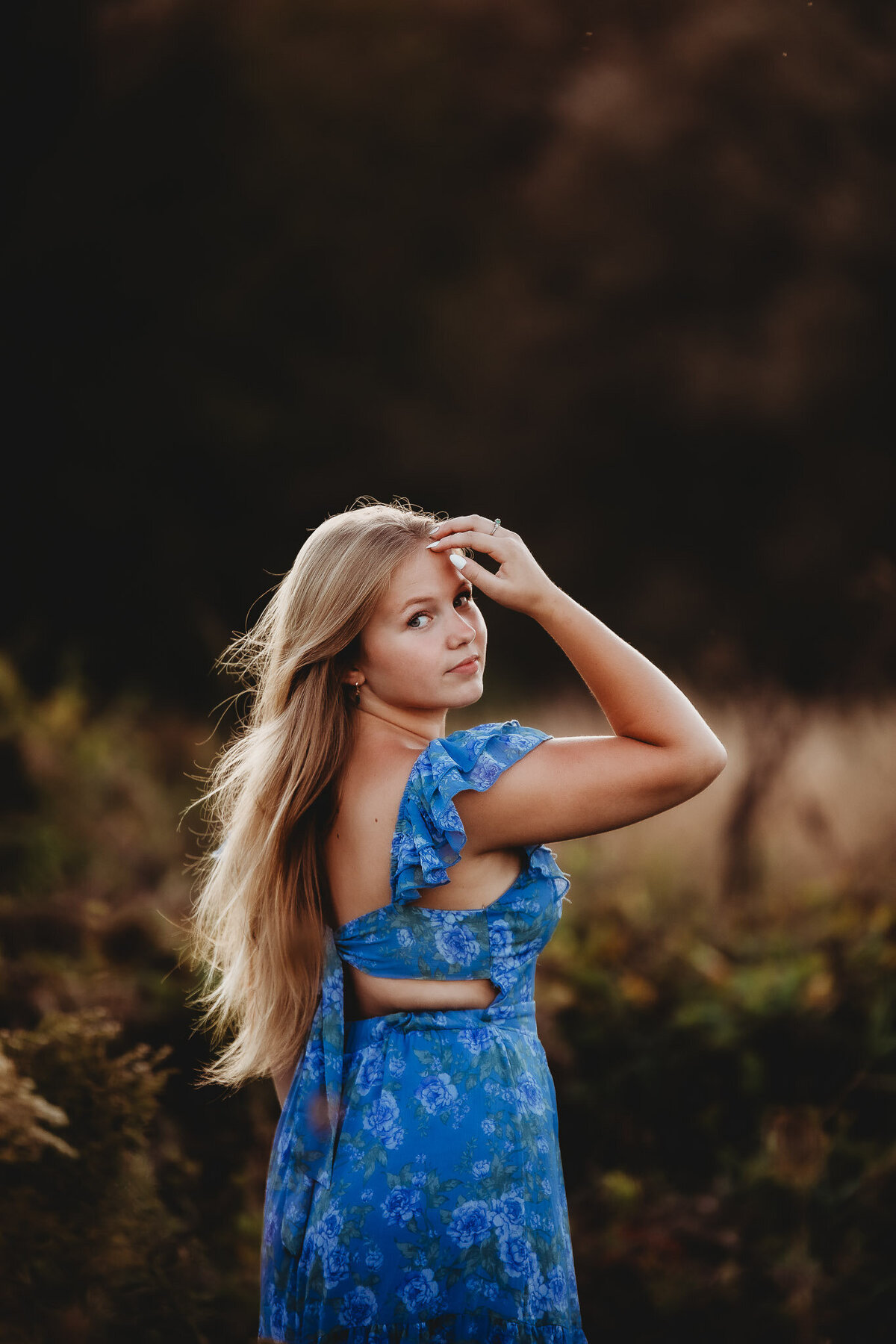 High school senior girl outdoors in a blue dress with her hand up by her forehead, long blond hair flowing in the wind and sunlight