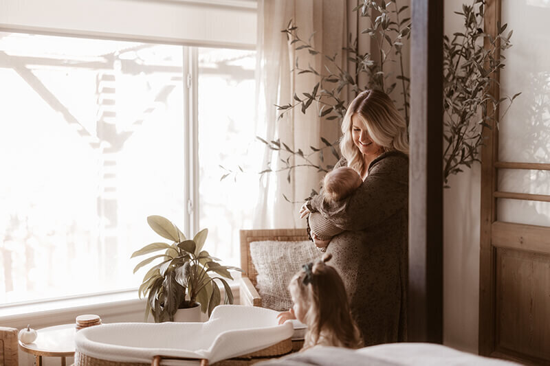 mom cuddles newborn baby boy while looking down at toddler daughter in master bedroom