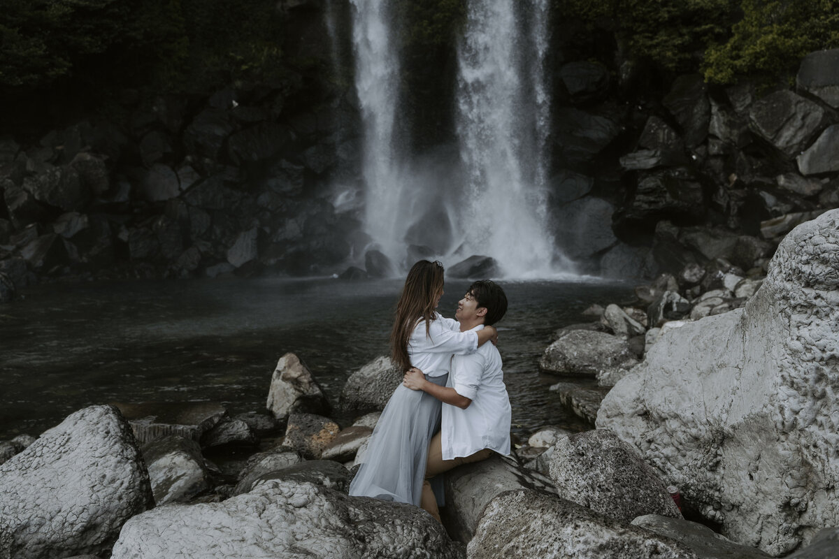 the groom sitting on the rock while holding the bride's waist facing each other in jeongbang waterfall