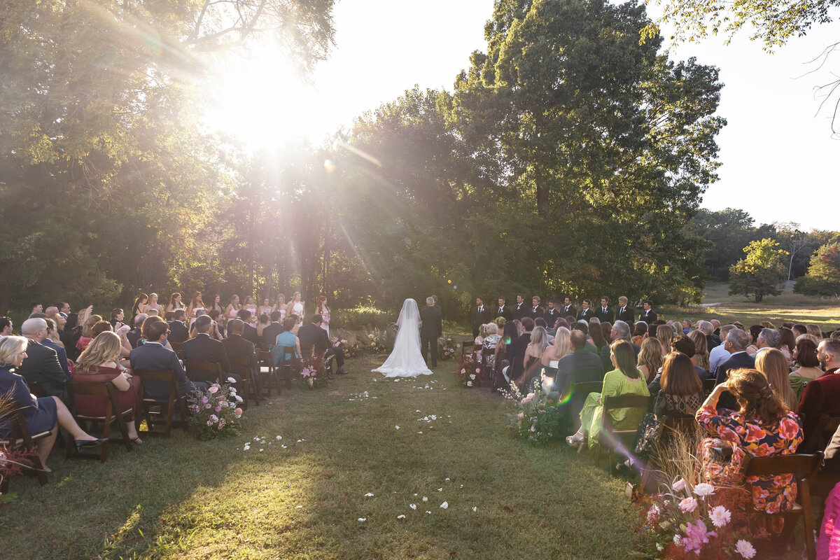 Outdoor wedding ceremony in a sunlit forest clearing with aisle flowers, seated guests, bridesmaids in blush, and a floral-accented arch framing the couple.
