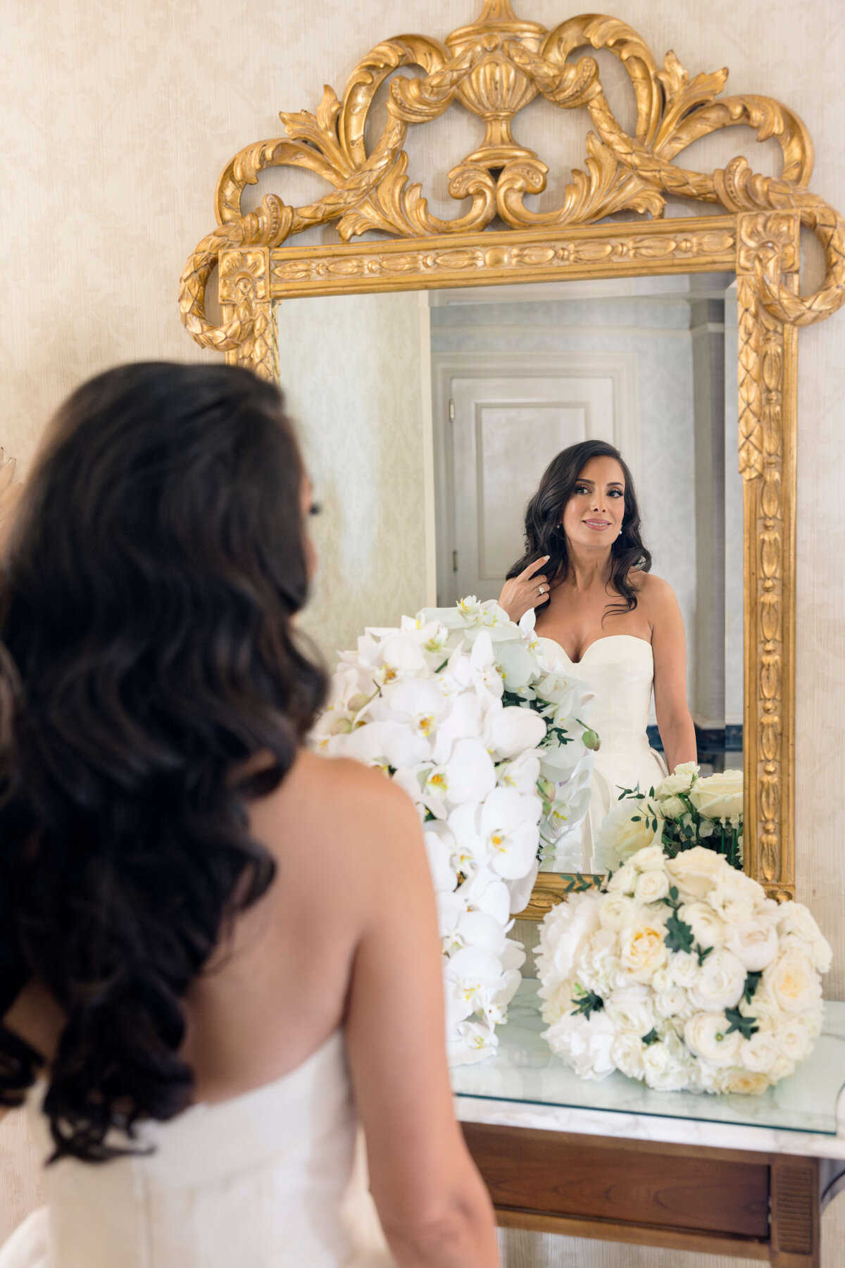 A woman in a white strapless dress stands in front of an ornate gold mirror, admiring her reflection. She is surrounded by white floral arrangements, and the room has an elegant, vintage style.