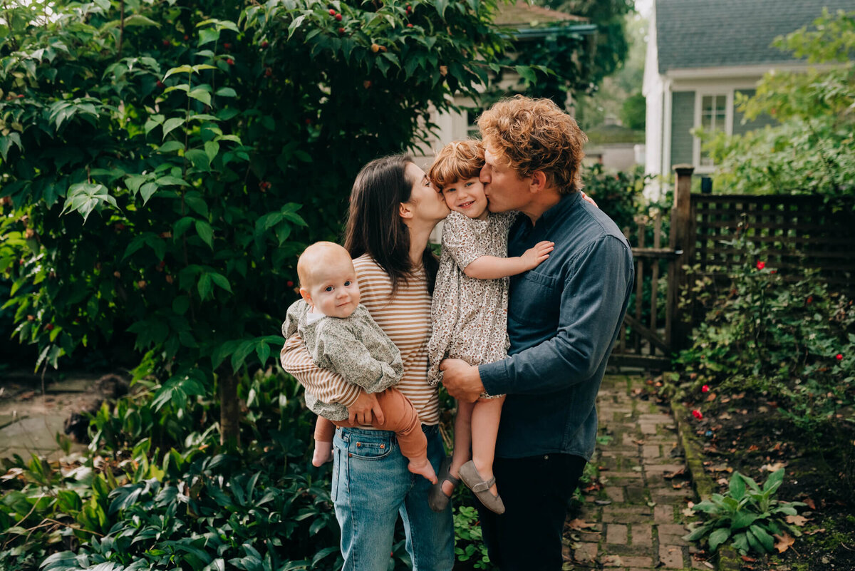 A mother and father hold and snuggle their two daughters in their back yard, both daughters are looking at the camera during their photoshoot with Boston Family Photographer, Allison Wolf