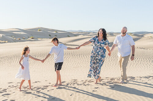 A mother, father, daughter, and son hold hands as they smile at each other while walking on the sand at Little Sahara recreation area.