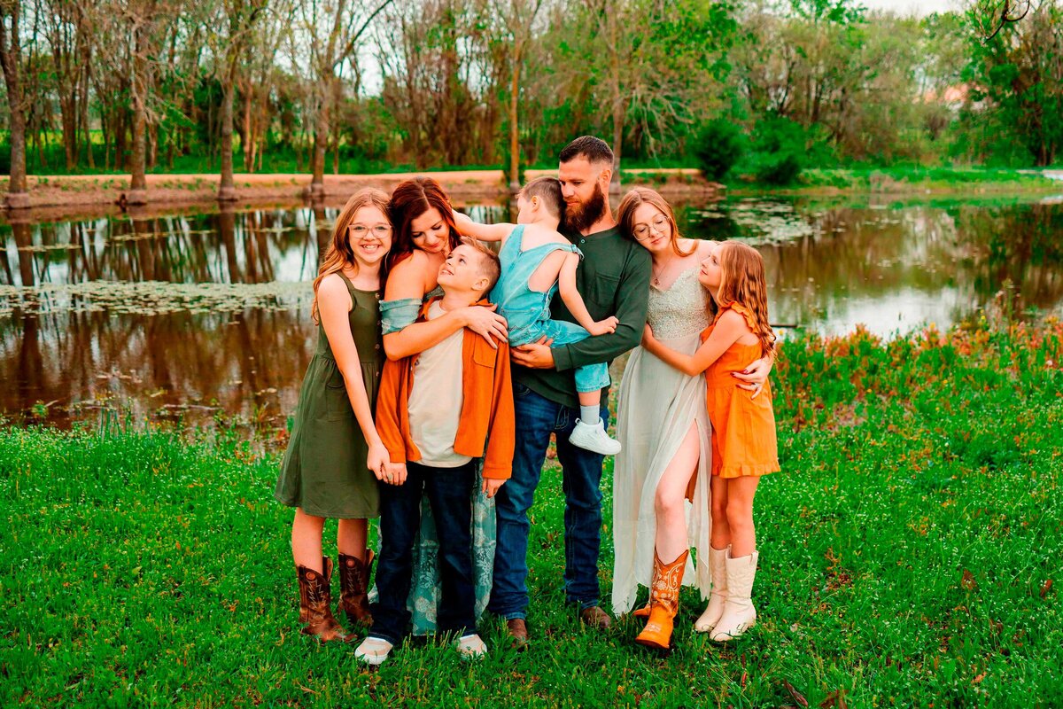 A joyful family poses in front of a serene lake with nature in the background. Dressed in vintage outfits, including long dresses, they exude happiness and contentment during their Albuquerque photography session.