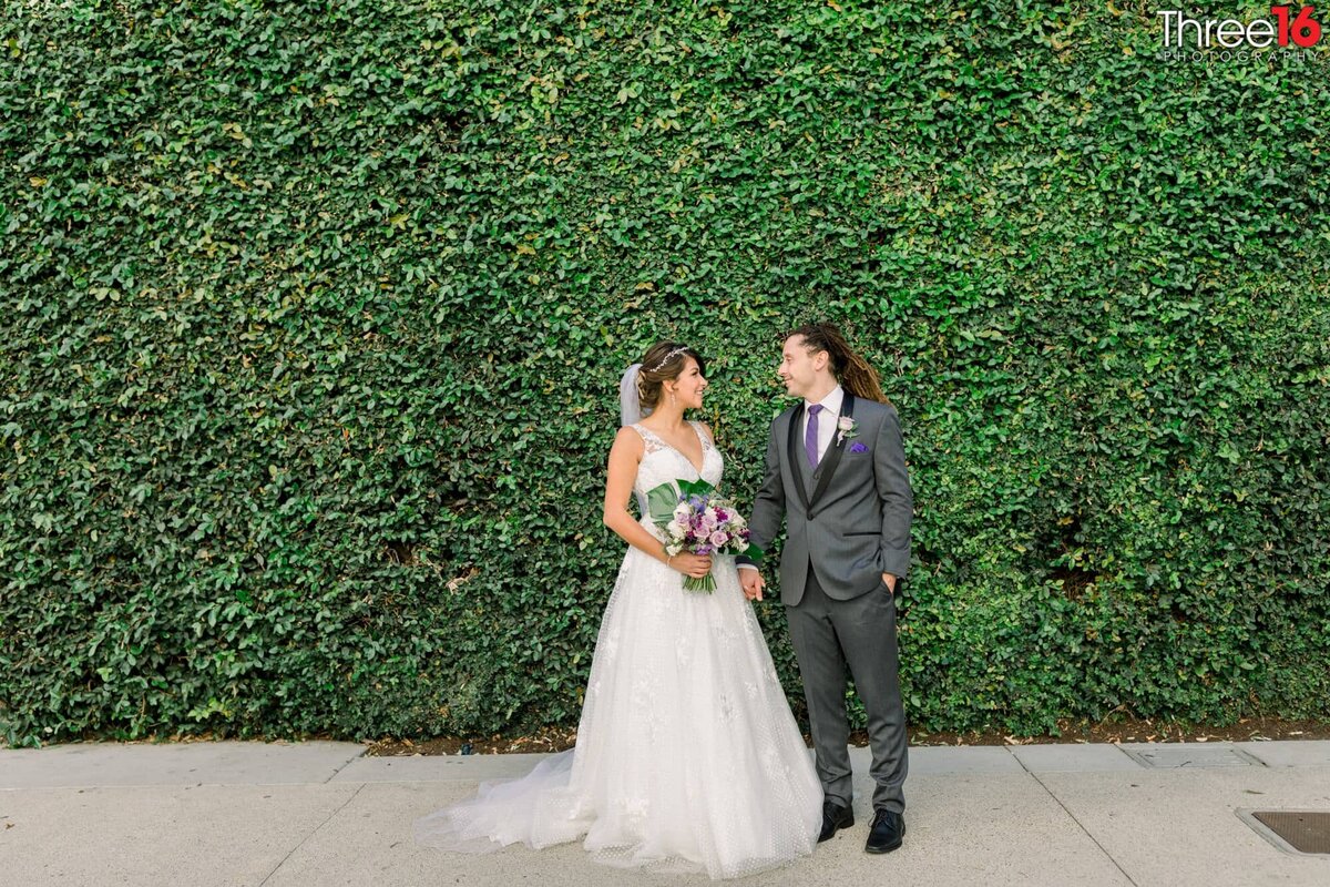 Bride and Groom smile at each other in front a of a ivy filled wall