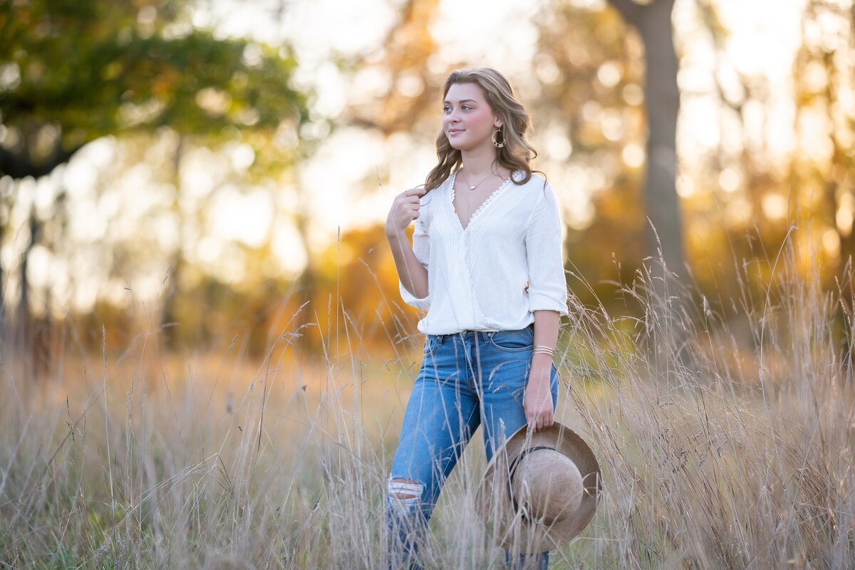 senior girl standing in dried grassy field holding hat