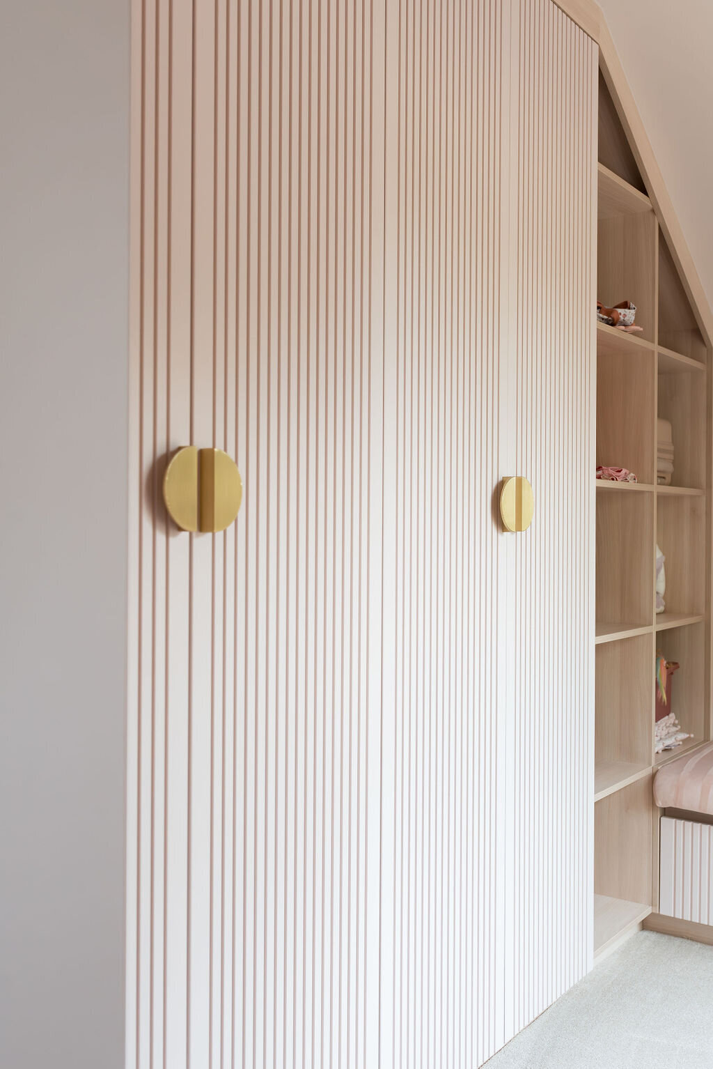 A modern pale pink wardrobe with vertically striped doors and large circular gold handles. To the right, there's an open shelf unit with various items and a bench with a cushion underneath.