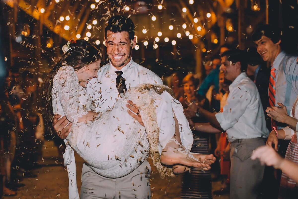 A bride and groom laugh amid a shower of confetti under twinkling string lights, capturing a jubilant wedding celebration moment.