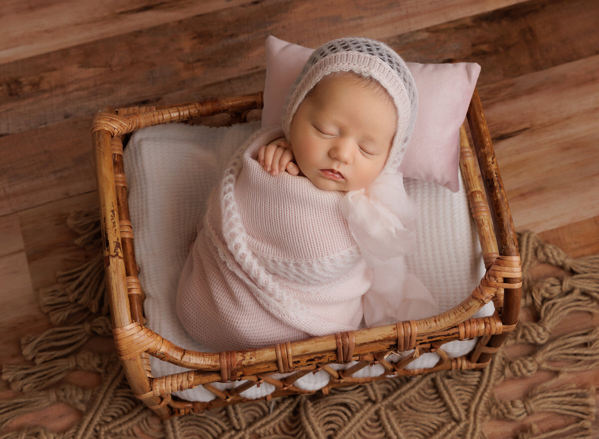 Baby girl in a pale blush swaddle is sleeping peacefully for this newborn photoshoot image.  Baby's hands are peeking out of the swaddle and she is wearing a matching bonnet. She is placed in a basket. Captured by top Brooklyn newborn photographer Chaya Bornstein