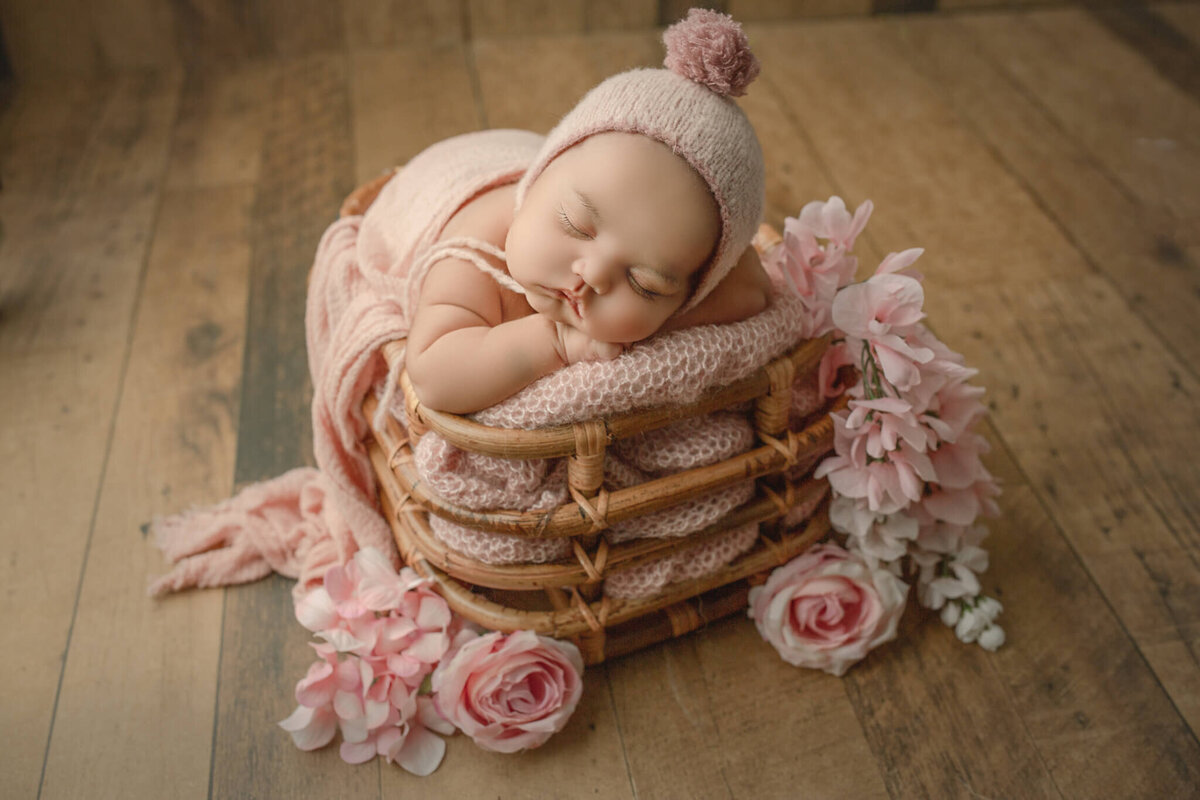 newborn baby girl poses with chin on hands in bamboo basket, surrounded by pink florals during her hamilton newborn photography shoot