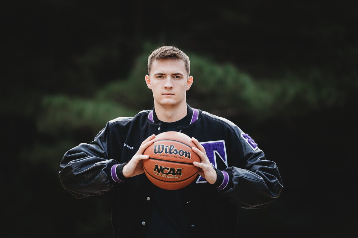 Dramatic senior portrait of a guy in his letter jacket holding his basketball in front of him.