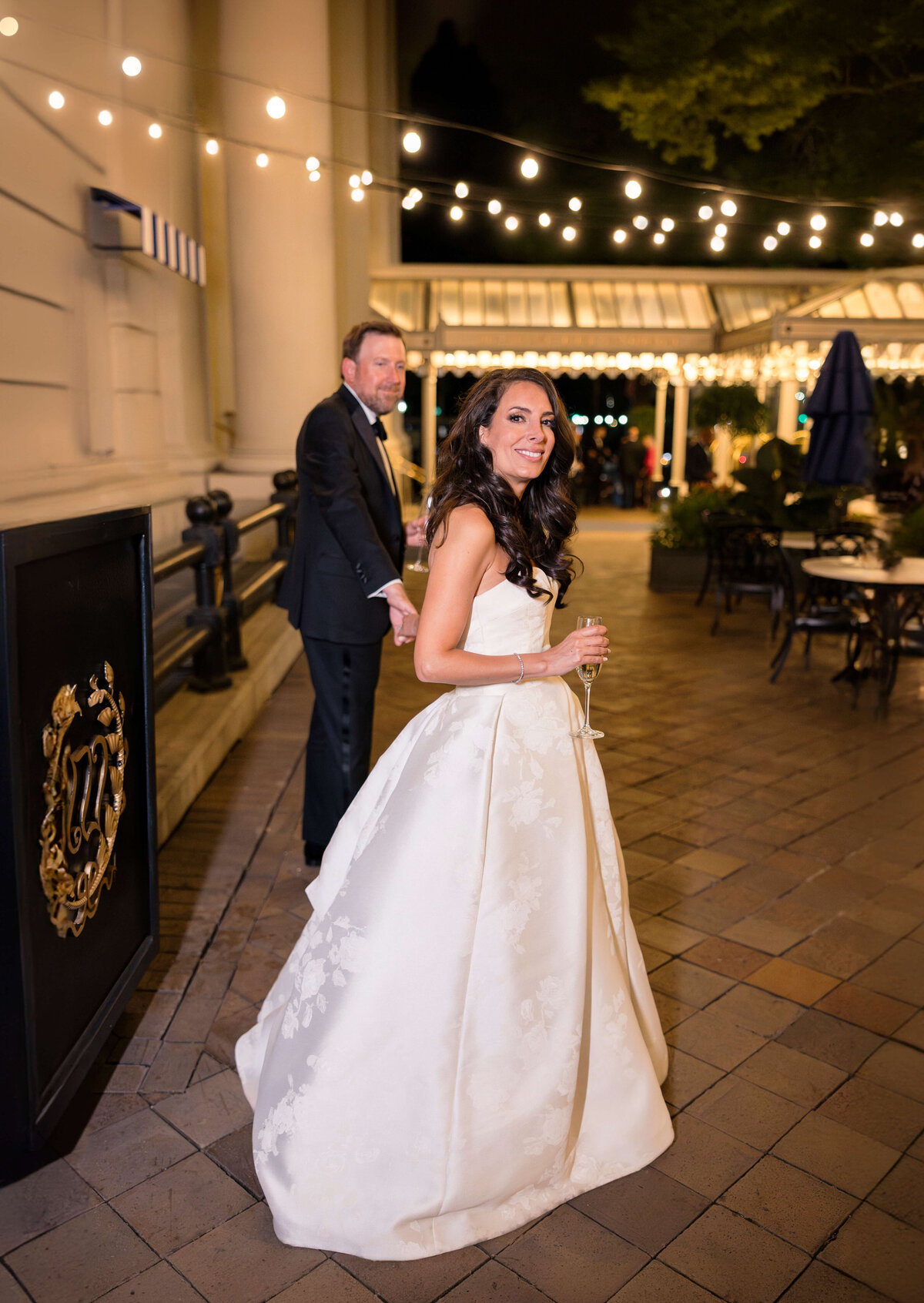 A bride in an elegant white gown smiles while holding a glass of champagne. A man in a suit stands behind her, also holding a glass. They are on an outdoor patio with string lights and ornate decorations.