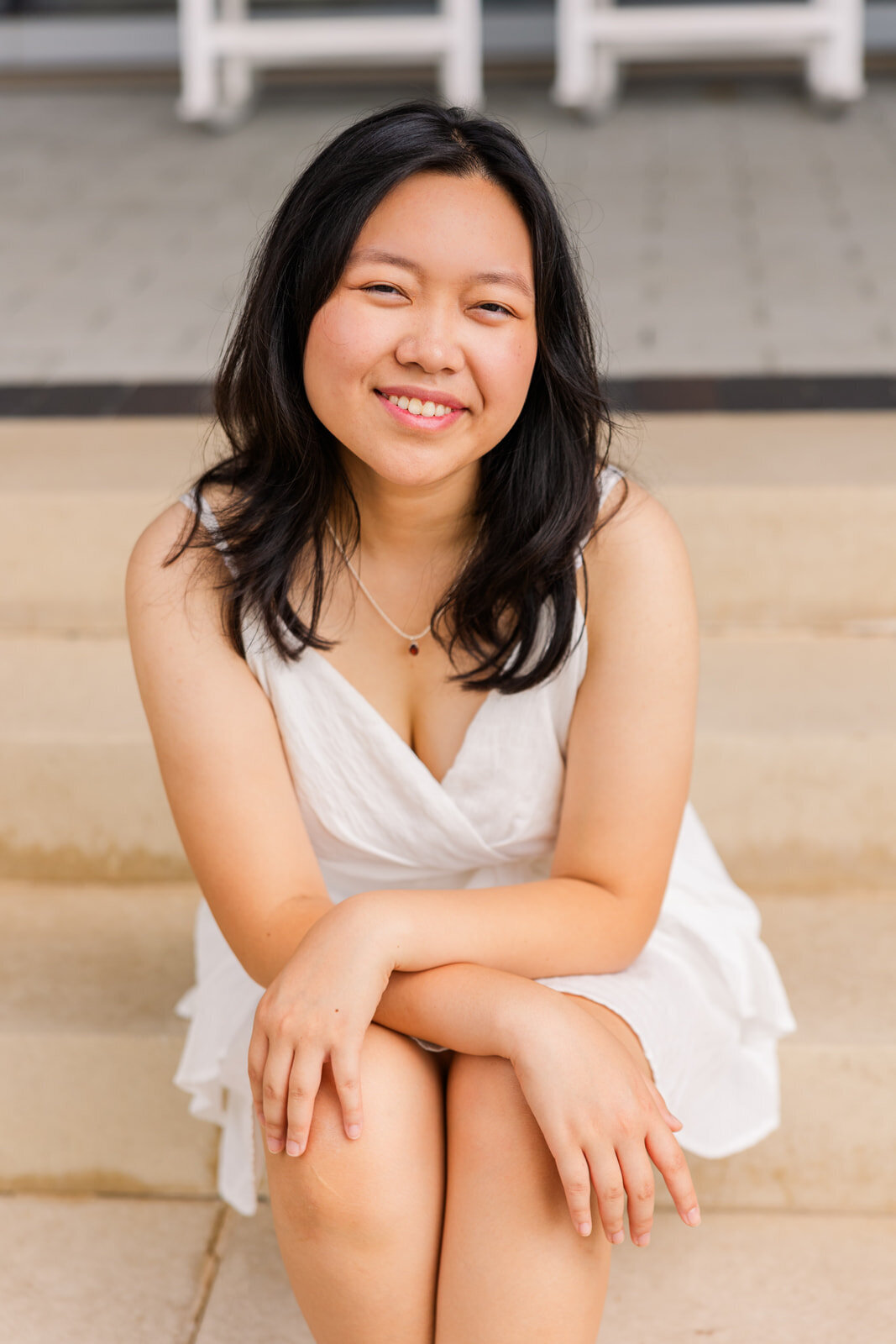 teenage girl wearing a white short dress sitting on stairs wrists crossed and hands resting on knees facing the camera during senior photoshoot with a neutral background