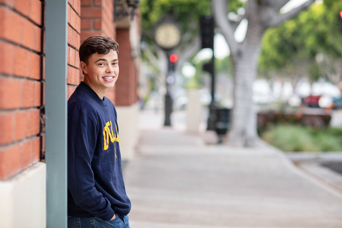 A person in a dark sweater leans against a brick wall on a sidewalk lined with trees and blurred street signs. The person is smiling, and the urban setting appears calm and pleasant.