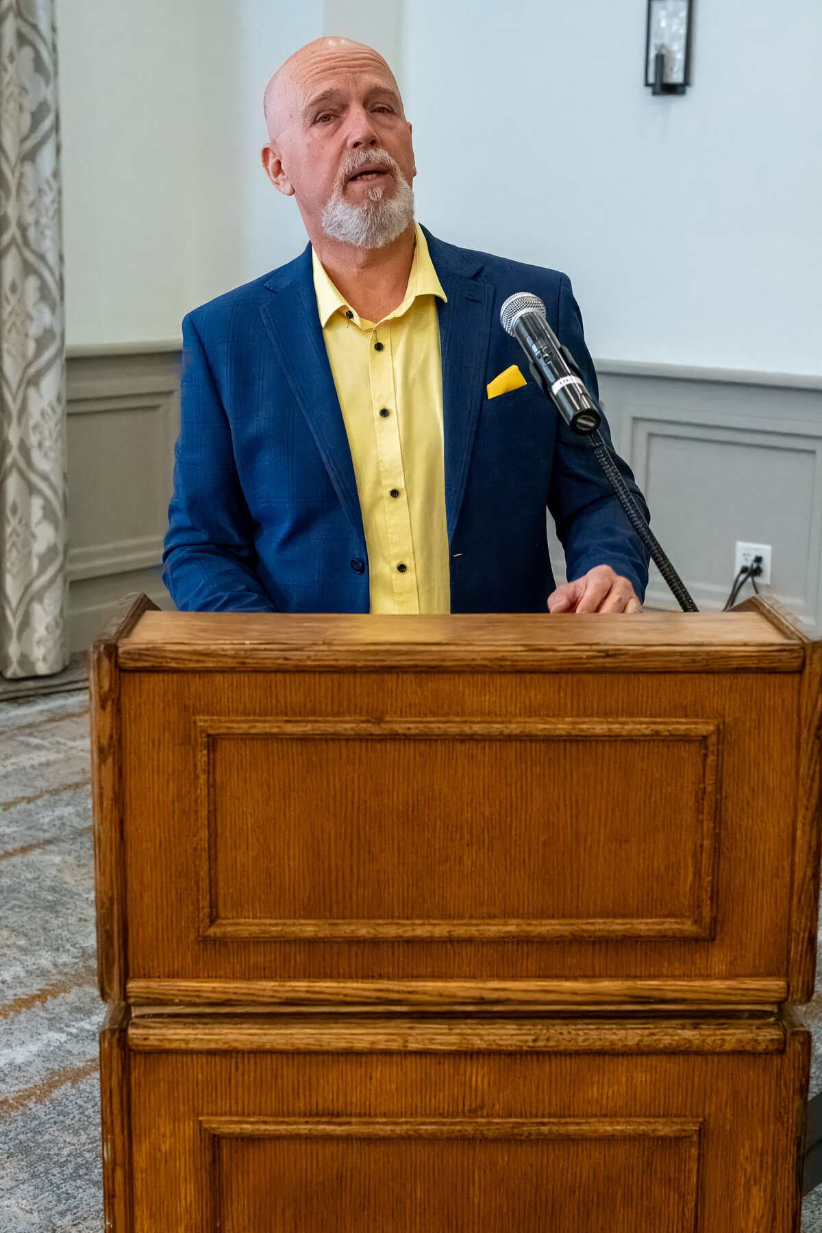 Man delivering a presentation behind a wooden podium, dressed in a blue jacket and yellow shirt, captured by Vyrl Photo for corporate branding.
