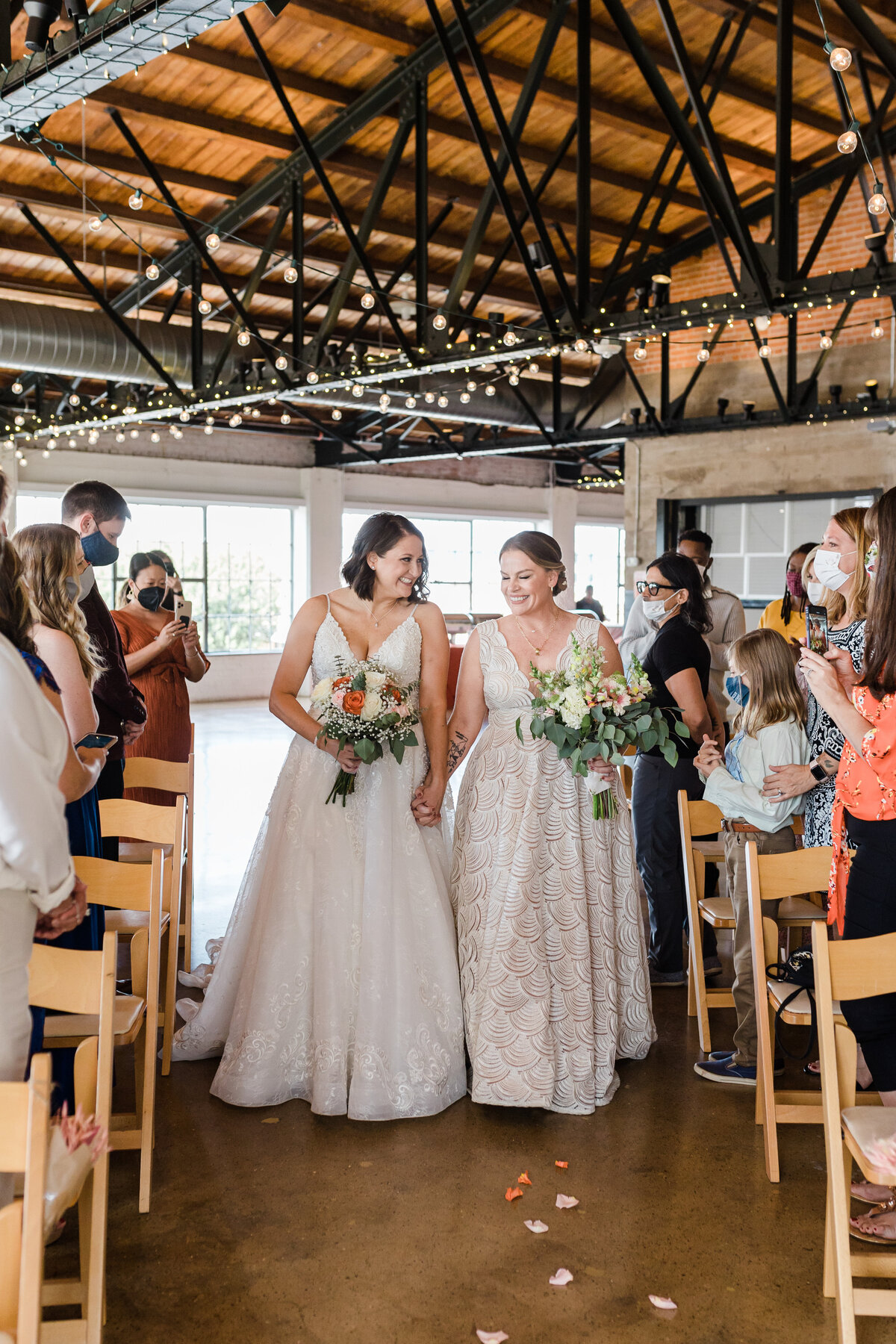 A candid shot of two brides walking down the aisle during their wedding ceremony at the Hickory Street Annex in Dallas, Texas. Both brides are wearing intricate, white dresses and are both holding bouquets. They are looking at each other and smiling joyfully. Many guests can be seen on either side of them.
