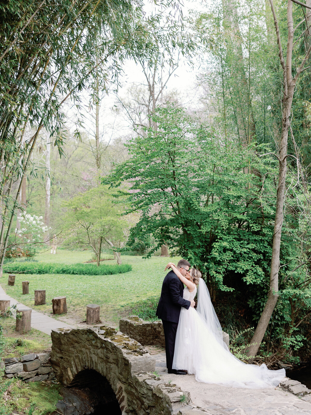 A bride and groom embrace and kiss on a stone bride with a small stream running underneath and tall tress in the background