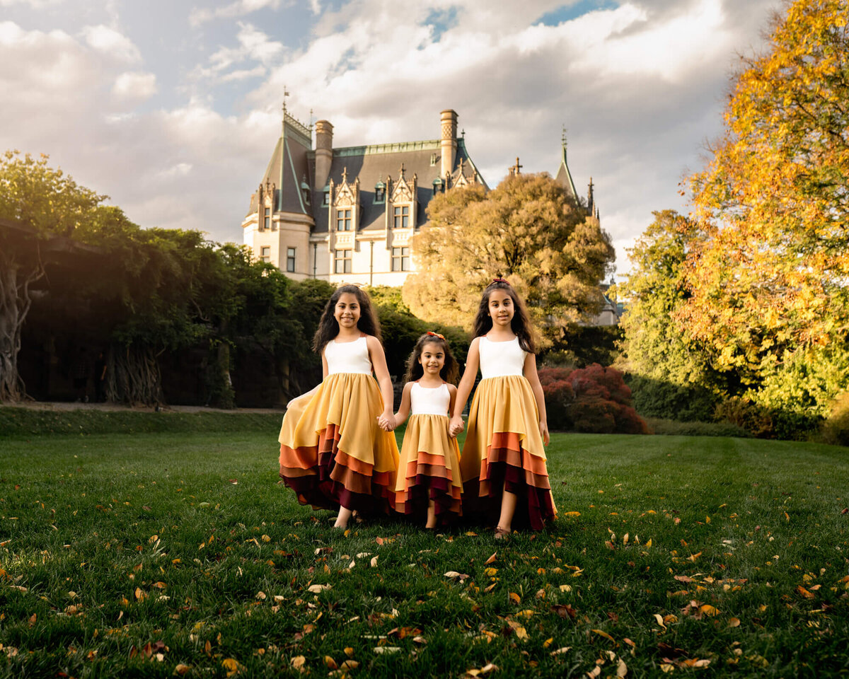 Three sisters dressed in long, beautiful dresses hold hands and walk in front of the Biltmore Estate