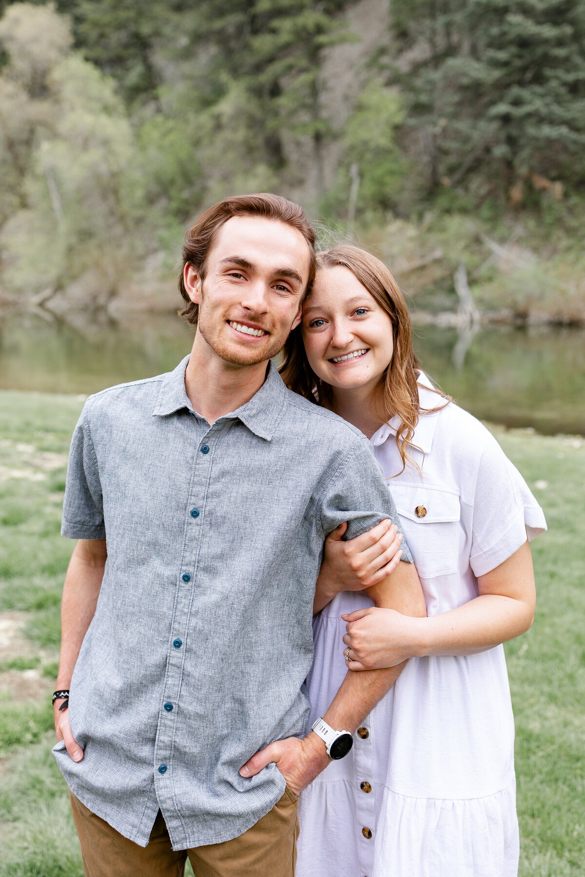 A woman rests her head on her fiancé’s shoulder, both smiling softly as they share an intimate moment by the river in Provo Canyon.