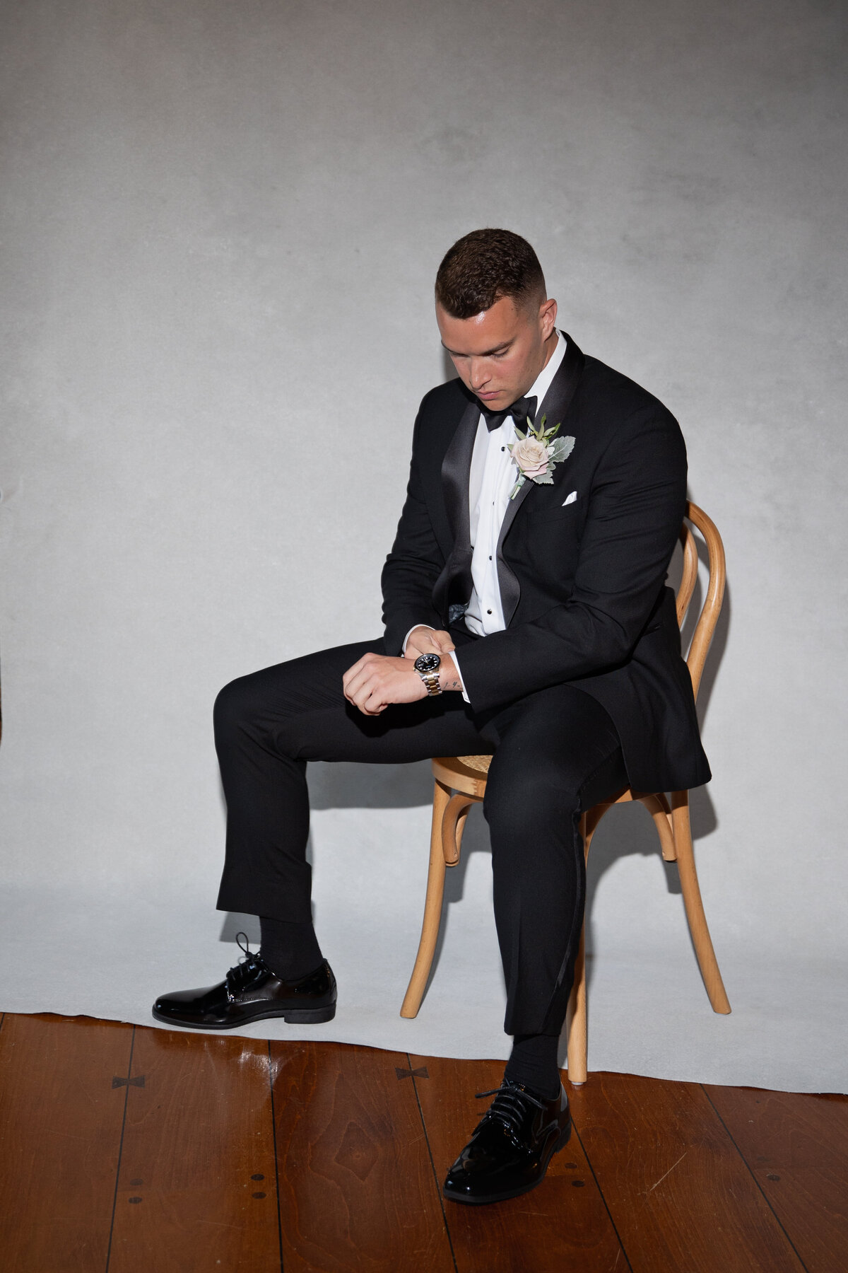 Aldrich Mansion Wedding Photos | Kelsey Sheehan Photography Timeless Rhode Island Weddings | A man in a black tuxedo with a white shirt and floral boutonniere sits on a wooden chair, looking at his watch. The setting is minimal and formal.